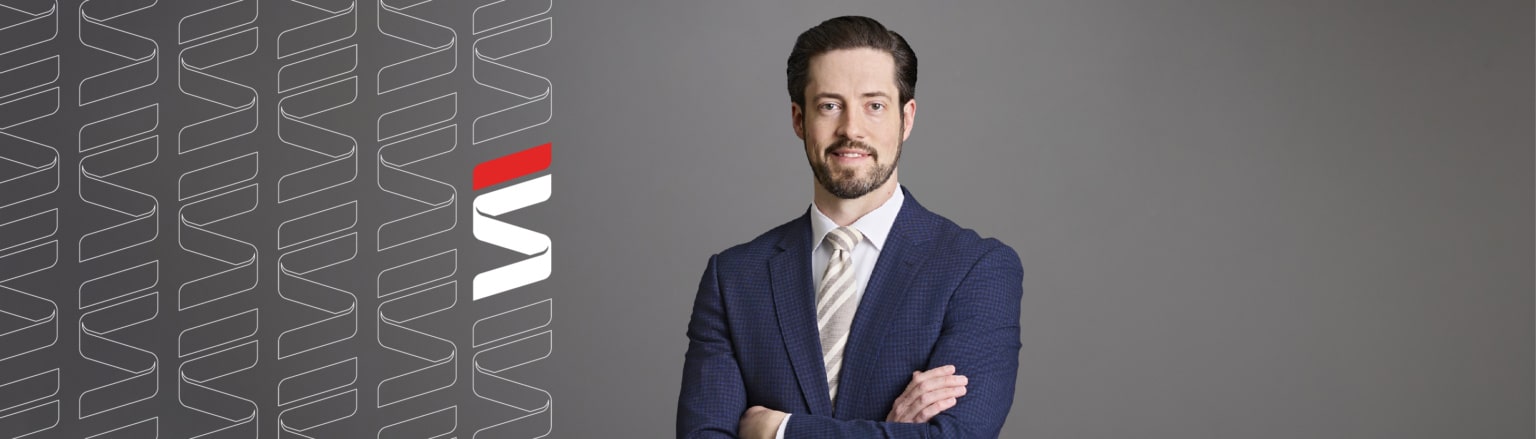 Sean Lavery, Fraser Stryker attorney, wearing a dark blue checkered suit jacket, white shirt button down shirt, and grey and tan striped tie. Fraser Stryker logo pattern on the left side.
