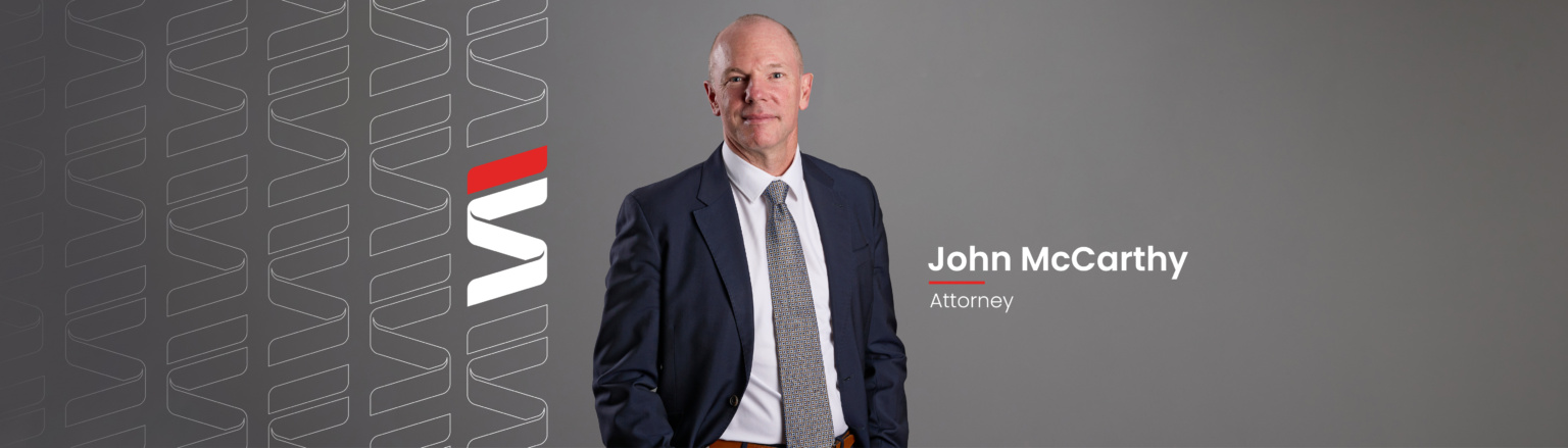 John McCarthy, Fraser Stryker attorney, wearing a dark blue suit, white shirt button down shirt, and blue patterned tie. Fraser Stryker logo pattern on the left side.