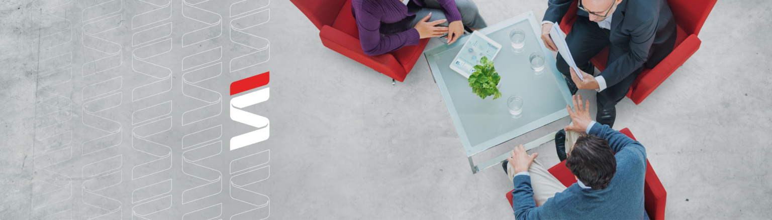 An overhead view of three people seated in red chairs around a small glass table in a modern setting, engaged in a professional conversation, with ample space on the light concrete floor surrounding them. Fraser Stryker logo icon repeating on the left side of the photo.